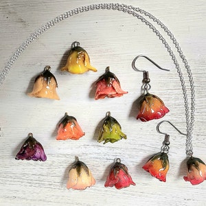 May include: A set of jewelry featuring delicate flower-shaped pendants and earrings. The necklace and earrings are made with a silver chain and feature a variety of colors, including yellow, orange, pink, and green.