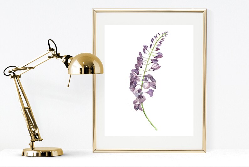 以下が含まれることがあります： 緑の葉がついた紫色の藤の花の茎の水彩画イラストが描かれた額入りアートプリント。細いゴールドのフレームに入れられ、丸みを帯びたシェードと調節可能なアームを備えたゴールドカラーの金属製デスクランプの隣に配置されています。