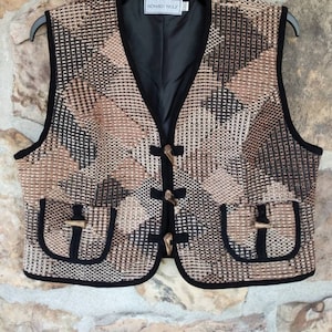 May include: A brown and black patterned vest with a black lining and a black trim. The vest has a V-neck and is fastened with three large buttons. The vest has two pockets with a flap and a button closure. The vest has a label that reads "Howard Wolf" on the front.
