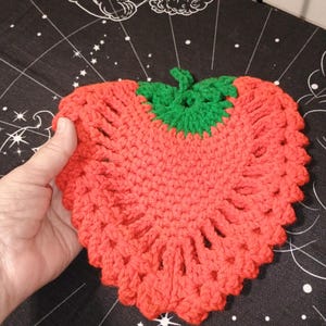 May include: A handmade, heart-shaped crochet item in vibrant red, resembling a strawberry. The top features a green stem. The item is held by a hand, showcasing the intricate stitch work. This decorative piece could be a coaster or trivet.