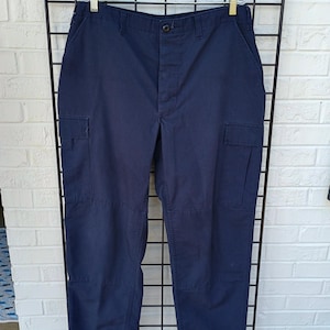 May include: Navy blue cargo pants featuring a button closure, a zippered fly, belt loops, and two large side cargo pockets with flaps. The pants have a straight-leg cut and are displayed on a clear plastic hanger against a black wire grid.