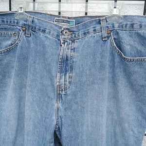 May include: Light blue denim jeans with a relaxed fit, featuring classic five-pocket styling. The jeans have a button and zipper closure, belt loops, and are hanging on a white plastic hanger.
