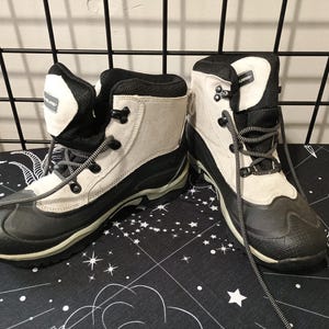 May include: Pair of black, white, and gray winter boots. The boots have a black rubber sole, a white upper, and black accents. They feature gray laces and are designed for outdoor use.