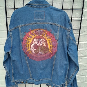 May include: A blue denim jacket featuring a large, circular graphic on the back. The design depicts a central face with three profiles surrounded by a stylized sun with red and yellow rays, intricate patterns, and a small waterfall landscape.