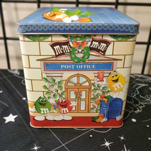May include: A rectangular tin box featuring a festive "Post Office" design with M&M's characters. The box has a blue roof with an orange M&M character and a brick facade. The front of the box has a door, a mailbox, and Christmas decorations.