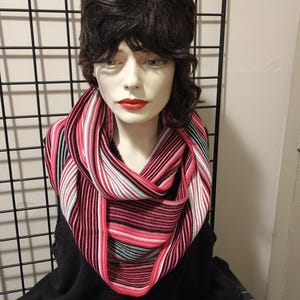 May include: A striped infinity scarf in shades of pink, black, white, and gray is draped around a mannequin's neck. The scarf's design features horizontal stripes, creating a bold and eye-catching pattern. The mannequin has dark hair and red lipstick.