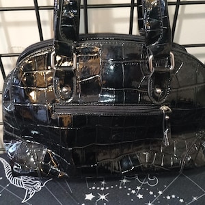 May include: A glossy black handbag with a crocodile skin texture. The bag has two handles, a zipper closure, and silver-toned hardware. The bag is sitting on a patterned surface.