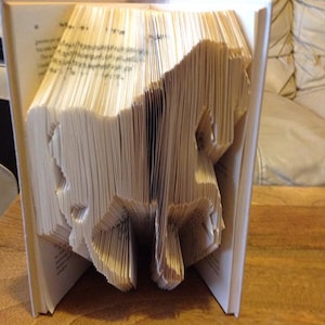 May include: A book sculpture of a horse, created by folding the pages to form the shape. The book is open, revealing the horse silhouette. The pages are a light cream colour, and the book is standing upright.
