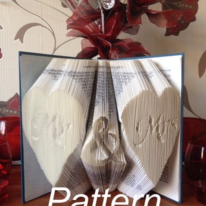 May include: An open book with folded pages, displaying heart-shaped cutouts with "Mr & Mrs" carved into the pages. The word "Pattern" is displayed at the bottom. The book is open, revealing the intricate design. A decorative floral arrangement is in the background.
