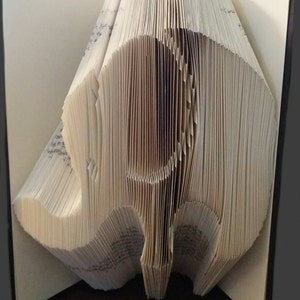 Pode incluir: Escultura de livro com páginas dobradas para criar a forma de um elefante. O elefante está em pé, com a tromba e as orelhas visíveis. As páginas são de cor bege claro, com as bordas ligeiramente mais escuras.