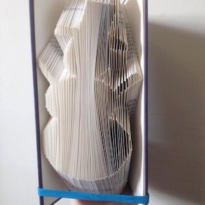 Peut inclure: Un livre dont les pages sont pliées en une sculpture 3D d'un arbre. Le livre est relié dans une couverture bleu foncé avec un ruban bleu maintenant les pages ensemble.