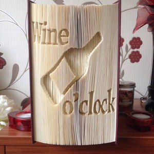 Könnte beinhalten: Ein Buch mit den Worten "Wine o'clock", die aus den Seiten ausgeschnitten sind. Das Buch steht aufrecht auf einer Holzoberfläche.