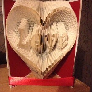 May include: A book folded into the shape of a heart with the word "LOVE" spelled out in the center. The heart is displayed on a red stand with a red ribbon around the base.