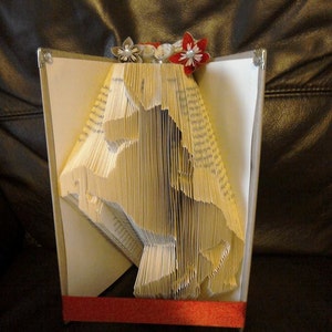 May include: A book folded into the shape of a unicorn, decorated with red, white, and gray paper flowers. The book is standing on a red base.
