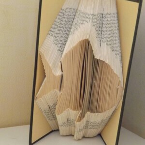 May include: A book art sculpture of a fish made by folding the pages of a book. The book is bound in black with yellow endpapers. The fish is made of folded pages and is standing upright.