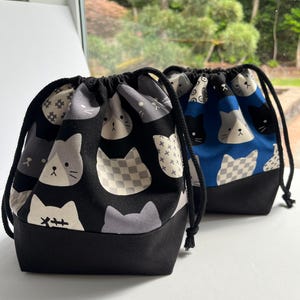 May include: Two drawstring pouches featuring a cat face pattern. The front pouch has a black background with gray, white, and checkered cat faces, while the back pouch has a blue background with similar cat faces. Both have solid black bases.