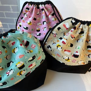 May include: Three drawstring bags with a sushi cat pattern. The bags have a black base and a patterned top with a black drawstring. One bag is light pink, one is light green, and one is beige.