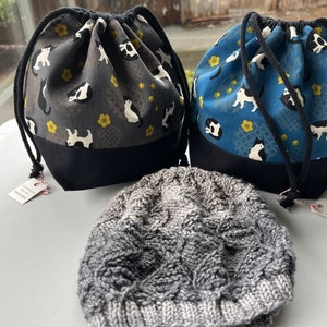 May include: Two drawstring bags with black and white cat designs on a blue and gray background. The bags are sitting on a white surface next to a gray knitted hat.