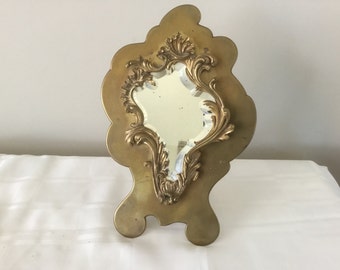 May include: An ornate gold-toned mirror with a decorative, asymmetrical frame. The mirror has a bevelled edge and is surrounded by intricate floral designs.