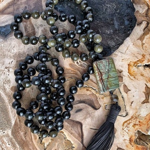 May include: A black and green beaded necklace with a tassel and a rectangular green and brown stone pendant. The necklace is lying on a wooden surface.