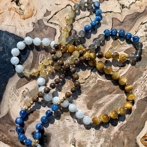 May include: A multi-strand mala necklace with a variety of gemstone beads, including blue, brown, gold, and white. The necklace has a large, gold-colored ammonite pendant.