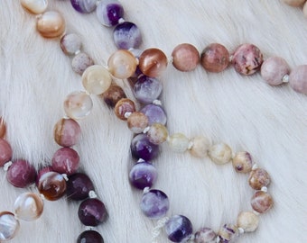 May include: A multi-colored gemstone mala necklace with a white tassel. The necklace features beads in shades of pink, purple, and brown.