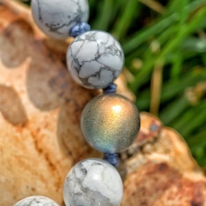 May include: A close-up of a white and gray beaded bracelet with a iridescent blue bead. The bracelet is on a wooden surface with green grass in the background.