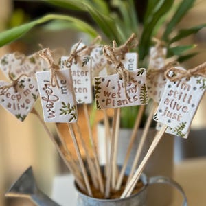 May include: A collection of white ceramic plant markers on wooden sticks, each tied with twine. The markers feature green leaf illustrations and text including "water me," "keep me alive," "i just wet my plants," and "i'm still alive. just."