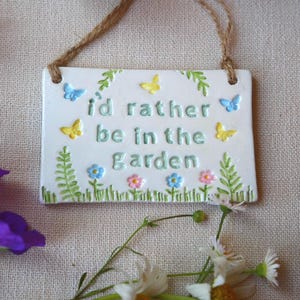 Op de afbeelding: Een rechthoekig wit keramieken bordje dat aan een jute touwtje hangt. Het bordje bevat de in reliëf aangebrachte groene tekst "id rather be in the garden", omringd door kleine geschilderde gele en blauwe vlinders, groene varens en bloemen.