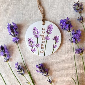May include: A circular white ceramic ornament featuring an embossed lavender plant design and the word Lavender in capital letters. The ornament hangs from a brown jute twine loop and is surrounded by several sprigs of fresh purple lavender.