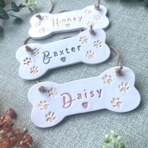 May include: Three white bone-shaped hanging ornaments featuring paw print engravings and small heart cutouts. Each ornament is personalised with the names Honey, Baxter, and Daisy in coloured lettering, suspended by thin brown twine strings.