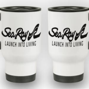 May include: Two white travel mugs with black handles and lids. The mugs feature the text "Sea Ray" in a stylized font above "LAUNCH INTO LIVING" in a sans-serif font. The mugs have a black base.