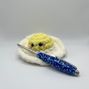 May include: A blue and white patterned pen with a silver tip and clip rests on a crocheted egg-shaped item. The egg has a yellow yolk and a white base. The pen has the text "F/5" on the silver tip.