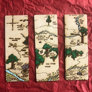 Könnte beinhalten: Drei Lesezeichen aus Holz mit einem Kartendesign, Text und Illustrationen. Die Karten zeigen Orte wie "Pooh Bear's House" und "100 Aker Wood" mit grünen Bäumen und blauen Flüssen. Die Lesezeichen sind rechteckig und hellbraun.