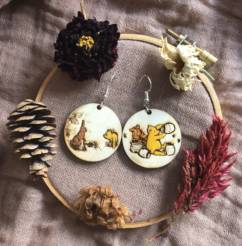 Puede incluir: Un par de pendientes de madera con los personajes de Winnie the Pooh y Piglet. Los pendientes son redondos y tienen un acabado de madera marr&oacute;n claro. Los personajes est&aacute;n representados en un estilo de dibujos animados y est&aacute;n pintados en amarillo, naranja y rojo.