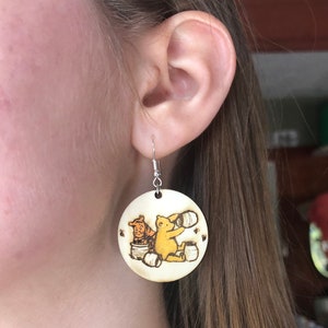 Puede incluir: Un par de pendientes de madera con un dise&ntilde;o de Winnie de Pooh y Tigger. Los pendientes son redondos y tienen un color madera claro. Los personajes est&aacute;n representados en un estilo de dibujos animados, con Winnie de Pooh sosteniendo un tarro de miel y Tigger sosteniendo un tarro de miel.
