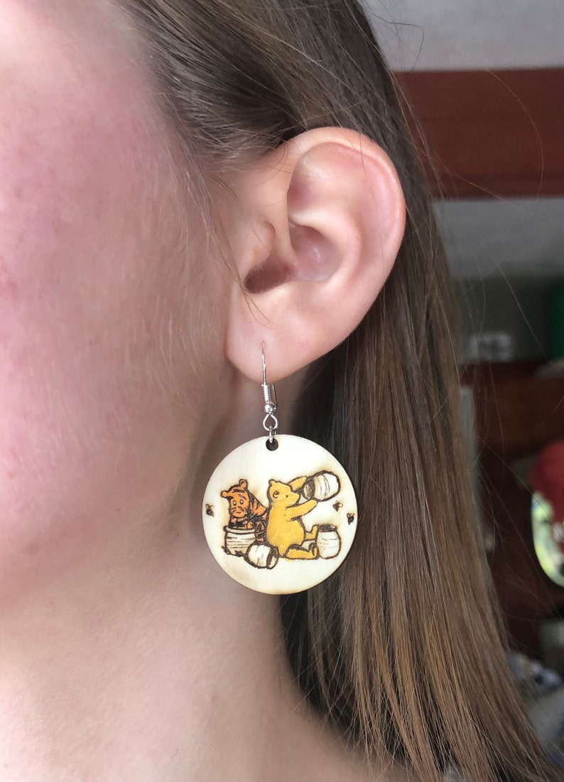 Puede incluir: Un par de pendientes de madera con un dise&ntilde;o de Winnie de Pooh y Tigger. Los pendientes son redondos y tienen un color madera claro. Los personajes est&aacute;n representados en un estilo de dibujos animados, con Winnie de Pooh sosteniendo un tarro de miel y Tigger sosteniendo un tarro de miel.