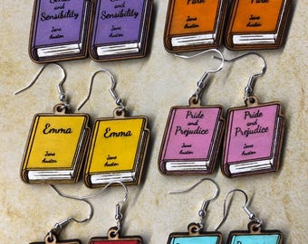 May include: A set of eight pairs of wooden earrings, each featuring a different Jane Austen novel cover. The covers are painted in various colors, including purple, orange, yellow, pink, red, and blue. Each earring features the title of the novel and "Jane Austen" in black text.