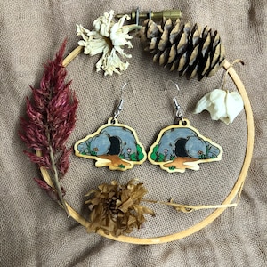 May include: A pair of wooden earrings with a cave design. The earrings are painted in shades of brown, gray, and green, with small white flowers around the cave entrance. The earrings are hanging from silver hooks.