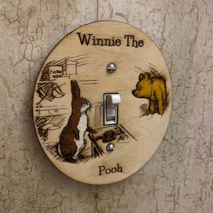 Pode incluir: Uma tampa de interruptor de luz redonda de madeira com uma ilustração de Winnie the Pooh. A tampa apresenta Winnie the Pooh e Porquinho do clássico livro infantil. O texto "Winnie the Pooh" está escrito na parte superior da tampa e "Pooh" está escrito na parte inferior.