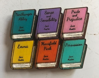 May include: Six wooden book-shaped ornaments featuring the titles of Jane Austen novels: Northanger Abbey, Sense and Sensibility, Pride and Prejudice, Emma, Mansfield Park, and Persuasion. Each ornament is painted in a different color and features the author's name.