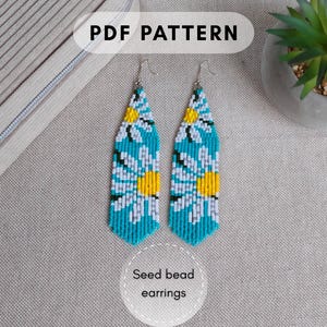 Puede incluir: Un par de pendientes largos de cuentas de semillas con forma puntiaguda y un patrón de margaritas en blanco, amarillo y azul cerceta. Tienen ganchos de metal plateado. La imagen incluye el texto PDF PATTERN y Seed bead earrings.