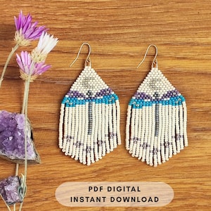 May include: A pair of handmade beaded earrings with a triangular design. The earrings feature a cream-colored base with accents of purple, turquoise, and gray beads. Long cream-colored fringe hangs from the bottom. The earrings are displayed on a wooden surface.
