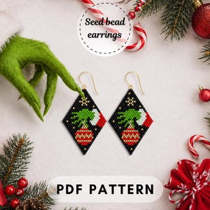 May include: A pair of diamond-shaped seed bead earrings featuring a black background with a green hand wearing a red and white cuff, holding a red and gold striped Christmas ornament. The earrings include gold-toned ear wires and a small snowflake.