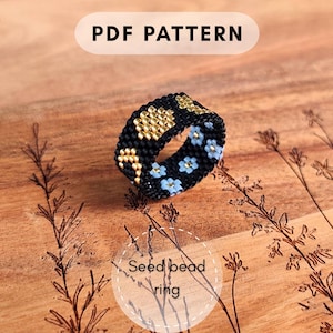 May include: A beaded ring with a black base, adorned with gold and light blue floral patterns. The ring is set against a wooden surface with etched floral designs. Text on the image reads "PDF PATTERN" and "Seed bead ring".