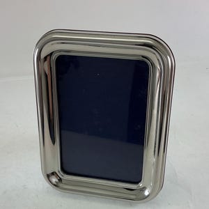 May include: A rectangular picture frame with a polished, reflective silver-tone metallic finish. The frame features a smooth, rounded border design with a deep, dark navy blue insert panel centered within the metallic structure.