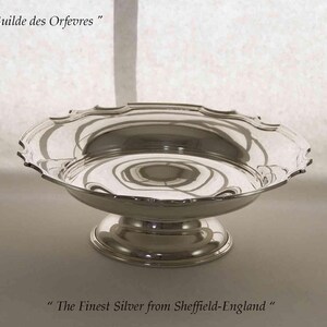 May include: A silver serving dish with a decorative, scalloped rim, supported by a pedestal base. The dish reflects light, and the text "Guilde des Orfevres" and "The Finest Silver from Sheffield-England" are visible.