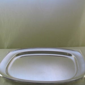 Puede incluir: Una bandeja de servicio rectangular de color plateado con esquinas redondeadas y una superficie metálica lisa y reflectante. La bandeja presenta un borde ligeramente elevado que enmarca el área central plana y pulida.