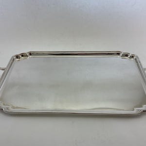 Op de afbeelding: Een rechthoekig zilverkleurig dienblad met een gepolijst, reflecterend oppervlak. Het dienblad heeft afgeronde hoeken met decoratieve inkepingen en twee stevige, geïntegreerde handgrepen aan de linker- en rechterkant voor gemakkelijk dragen.