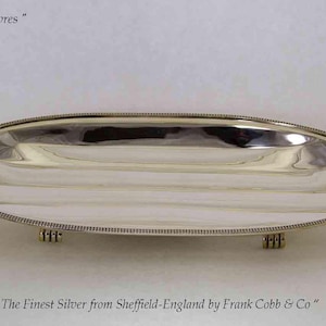 May include: A polished silver oval serving tray with a beaded border and small feet. The tray reflects light, with the text "Guilde des Orfevres" and "The Finest Silver from Sheffield-England by Frank Cobb & Co" visible.